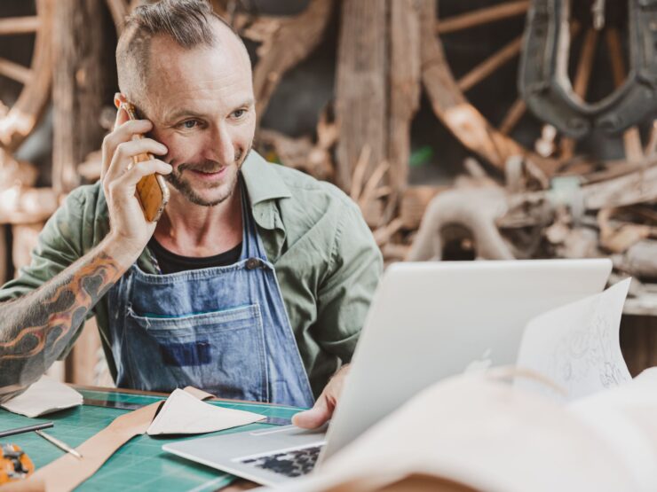 Mann in einer Werkstatt schaut auf einen Laptop und telefoniert mit einem Handy