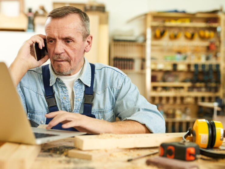 Mann arbeitet in einer Werkstatt und telefoniert an einem Handy und schaut auf einen Laptop